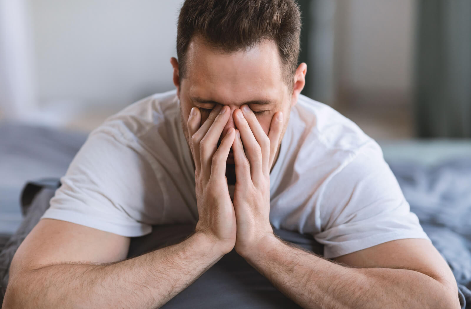 A man sitting on the side of his bed is massaging his irritated dry eyes.