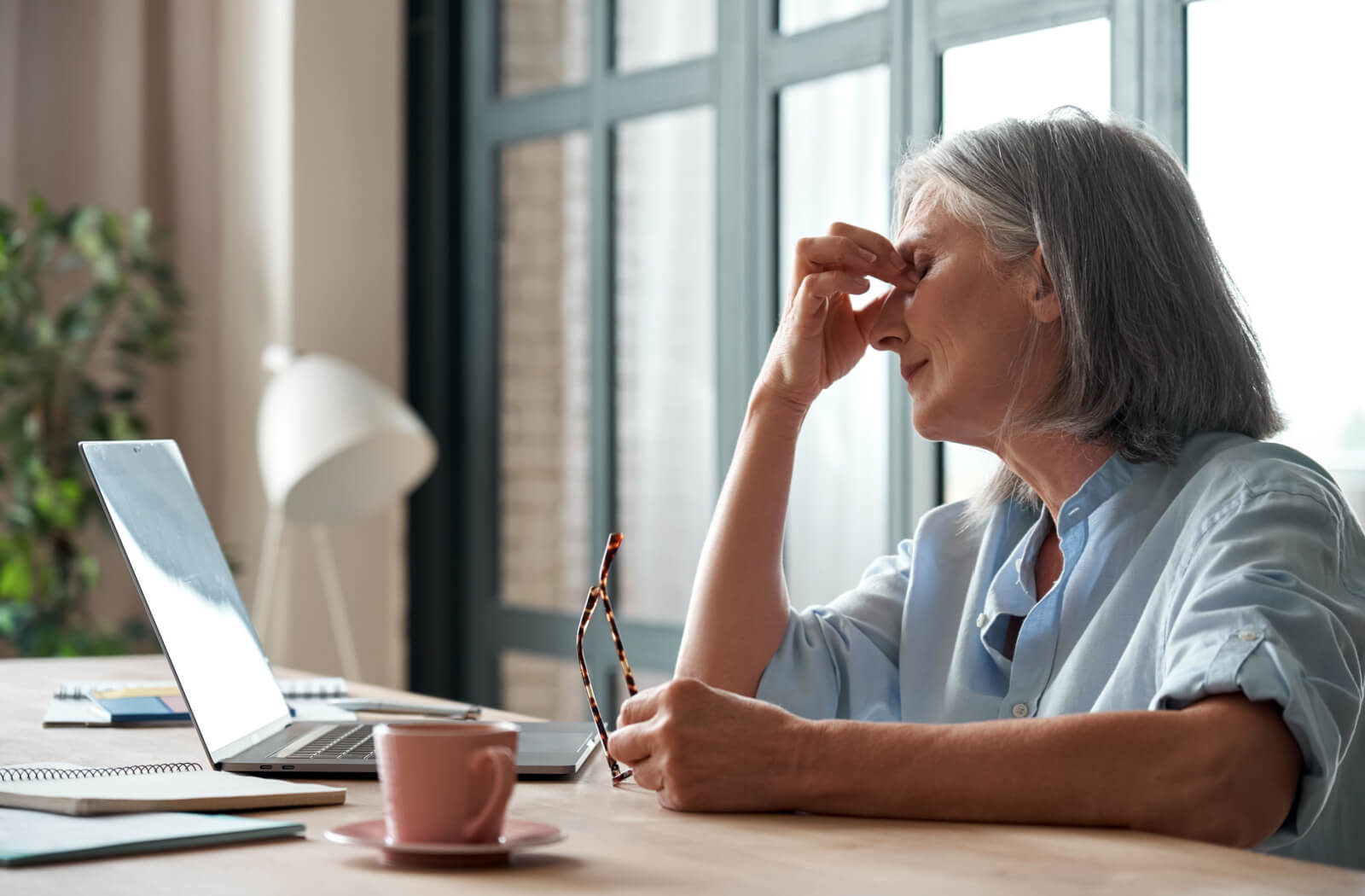 A senior woman sitting in front of her laptop computer takes off her eyeglasses and is suffering from dry eyes and a headache.