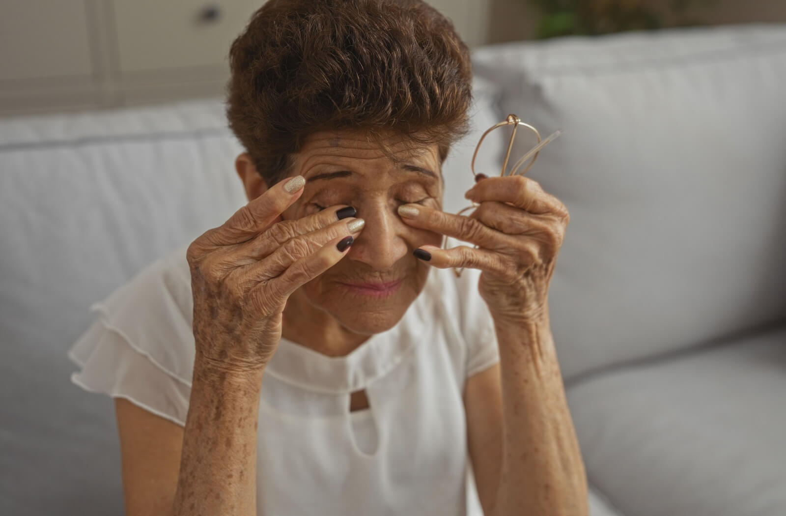 An elderly woman wearing a white shirt rubs her dry eyes with both her hands in discomfort.