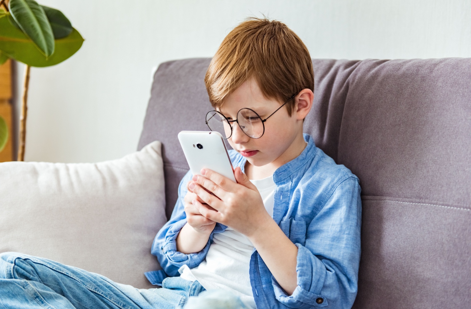 A child with myopia sitting on the couch, holding a cell phone close to their face and squinting.