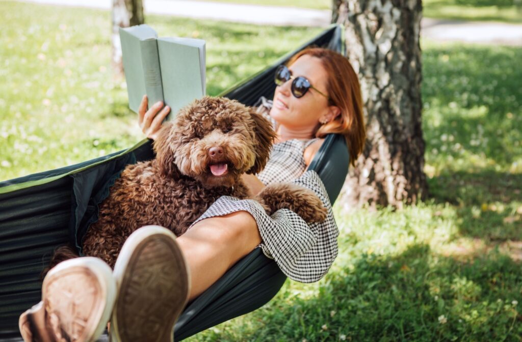 A woman relaxes in a hammock with their dog, able to easily read a book thanks to their prescription sunglasses.