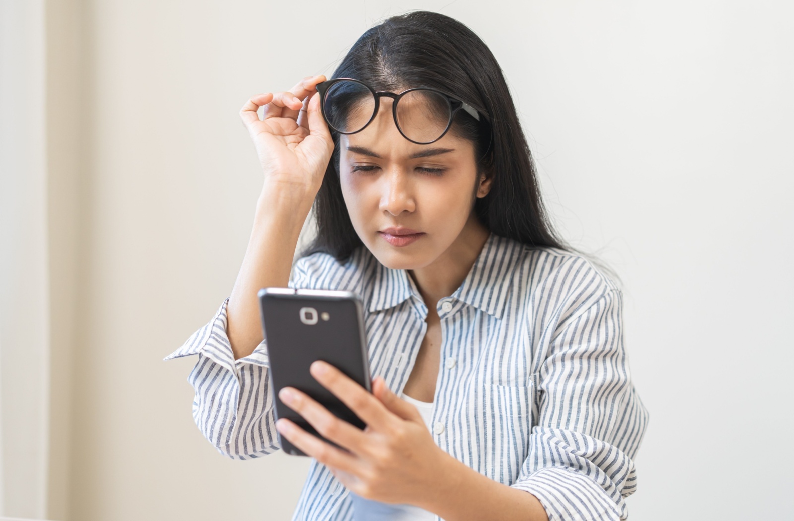 A woman lifts up their glasses and squints at their phone screen.