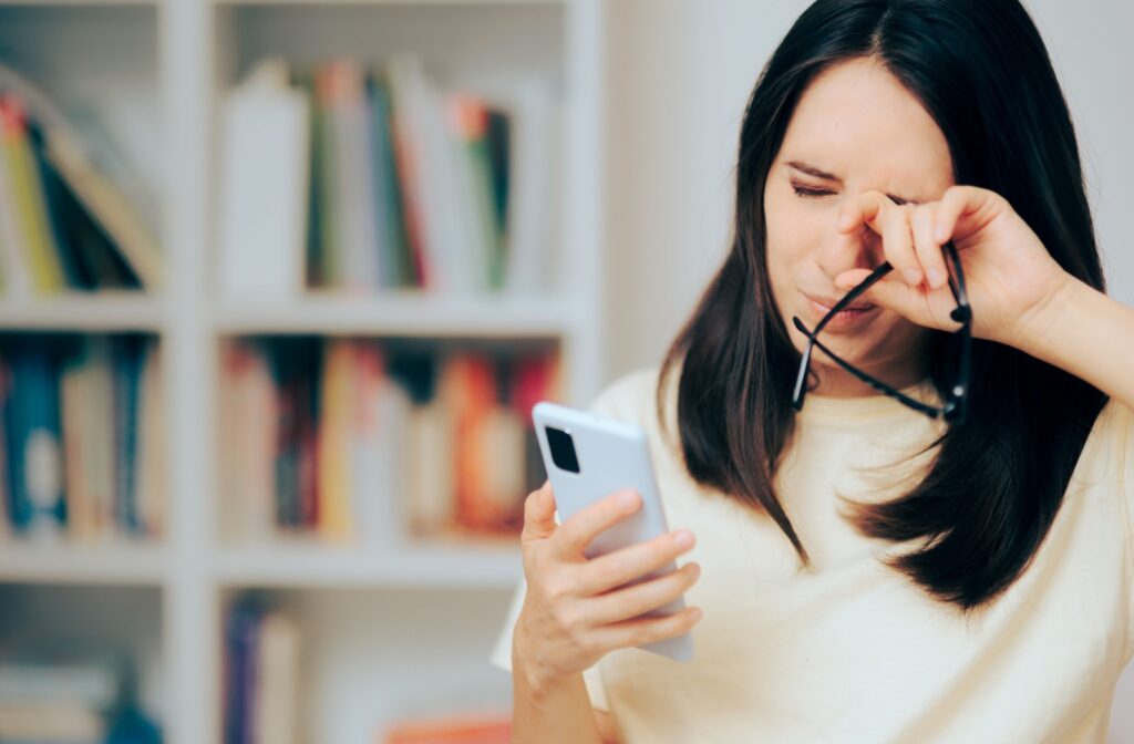 A woman rubs her eyes while holding her phone and glasses, showing discomfort from eye strain or dry eye symptoms.