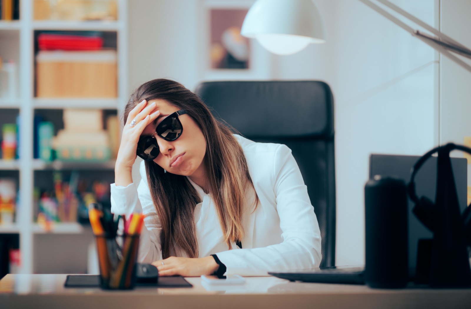 A woman sits at her desk wearing sunglasses indoors, resting her head in her hand due to light sensitivity and eye discomfort.