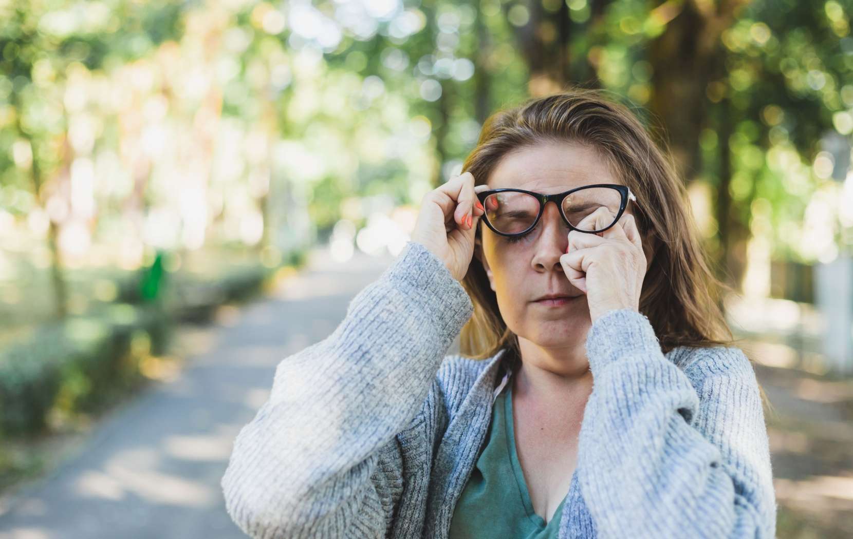 A woman standing outside lifts their glasses with one hand and rubs their tired eyes with the other hand.