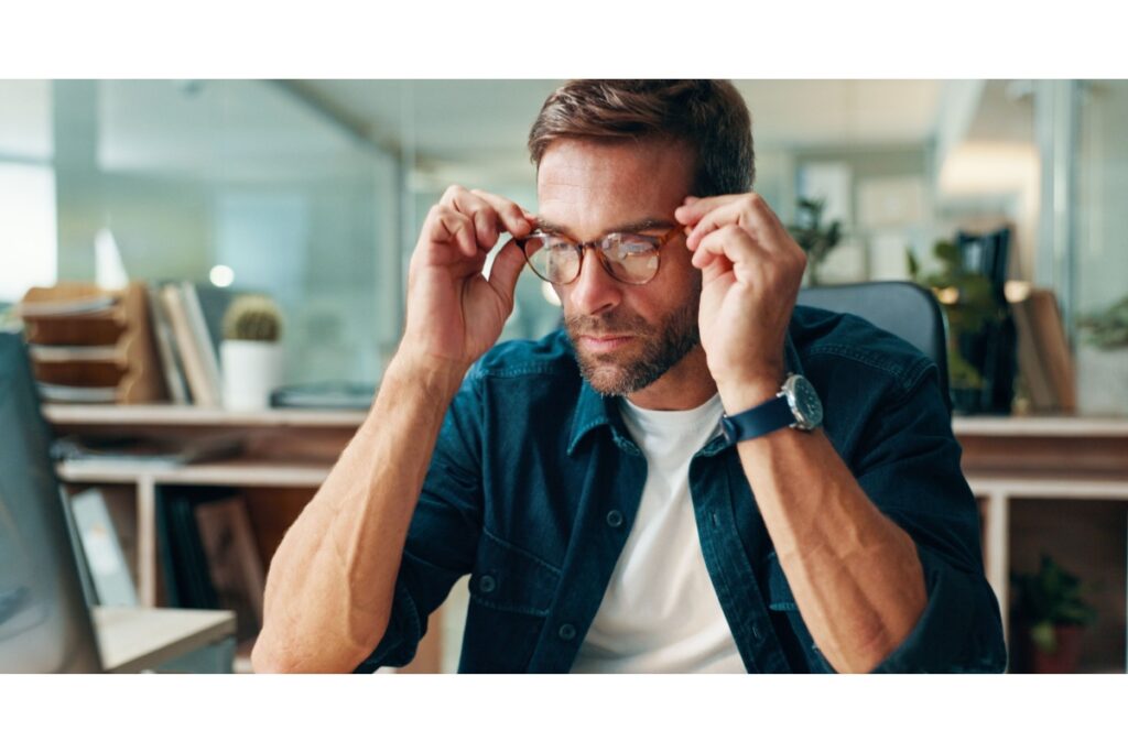 Middle-aged man in his 40s adjusting progressive eyeglasses while working at desk in home or office, demonstrating presbyopia and the need for multifocal lenses for clear vision at multiple distances.