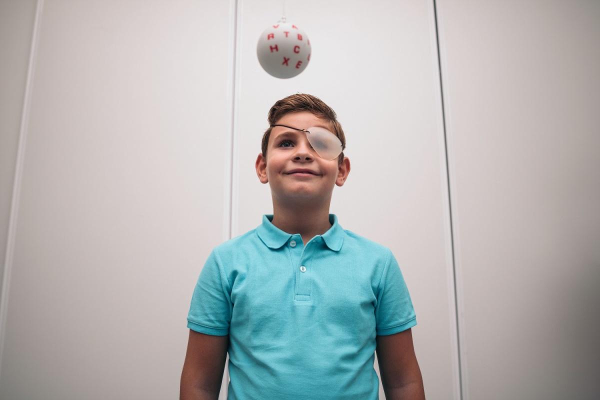 A smiling young boy wearing an eye patch performs a vision therapy exercise, looking up at a hanging ball with letters as he practices visual tracking and focus.