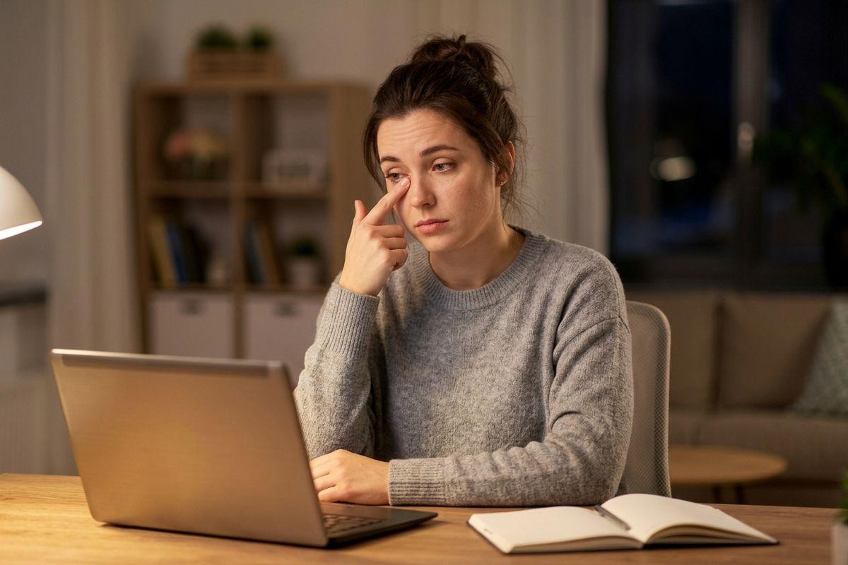 Adult sitting at a dimly lit home desk with a laptop, rubbing one eye with a finger, a notebook nearby.